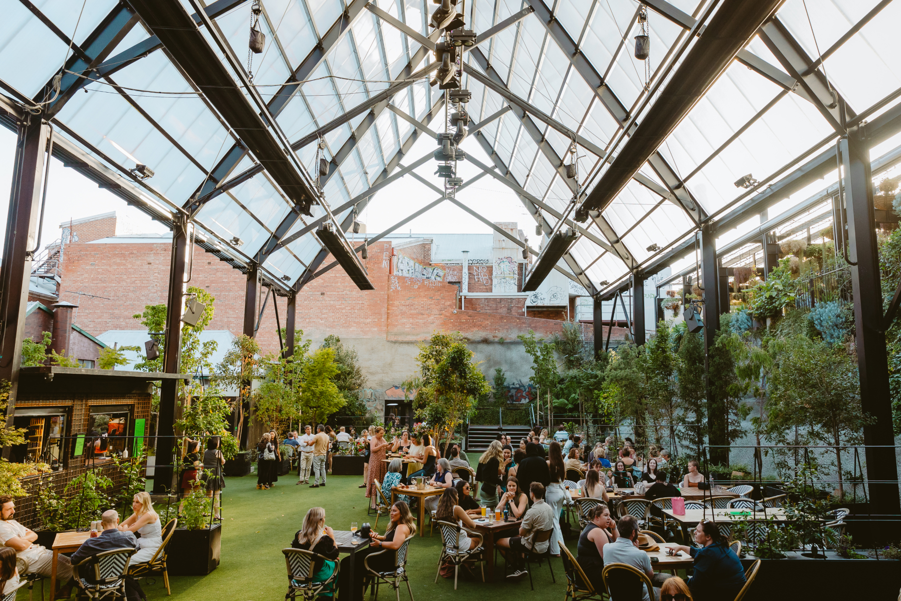 People gather and socialize at tables under a large, open-roofed greenhouse structure with glass panels, surrounded by plants and exposed brick walls, creating a lively outdoor dining atmosphere.