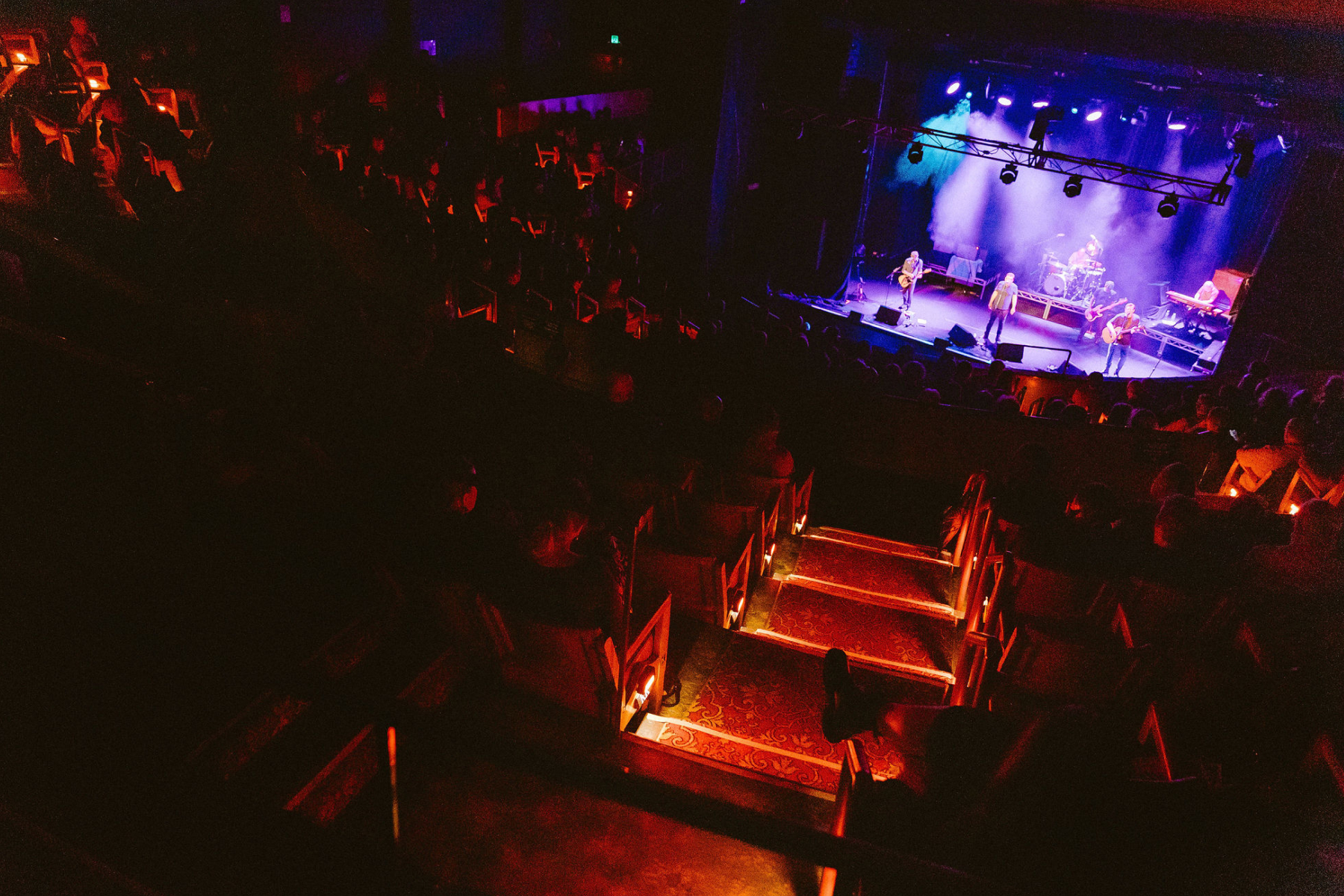 A dimly lit concert hall with glowing aisle lights, showing a band performing on stage under blue and purple lights while a seated audience watches from the darkened theater.