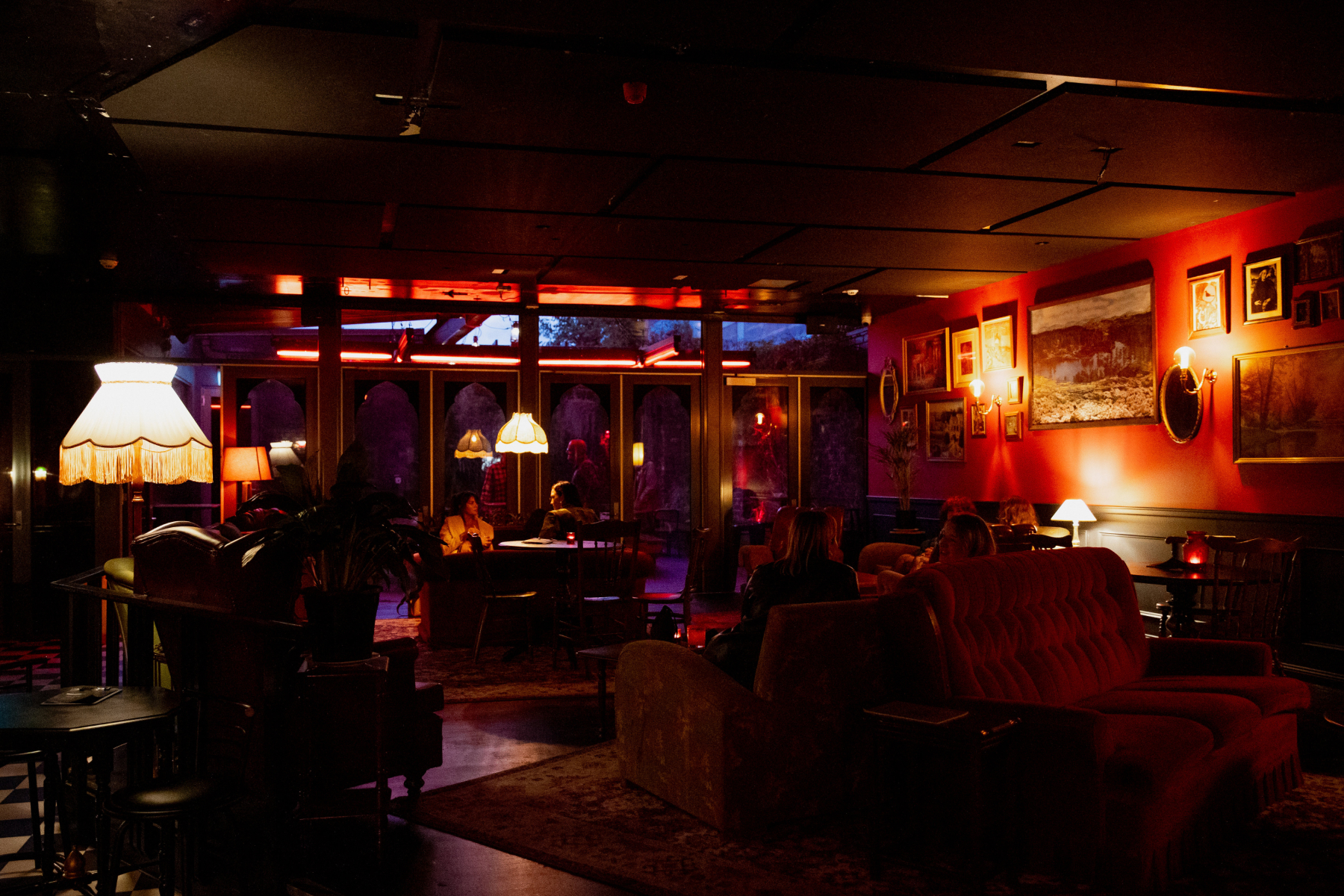 A dimly lit lounge with vintage lamps, red walls, framed pictures, cozy couches, and a few people seated, creating a warm, intimate atmosphere.